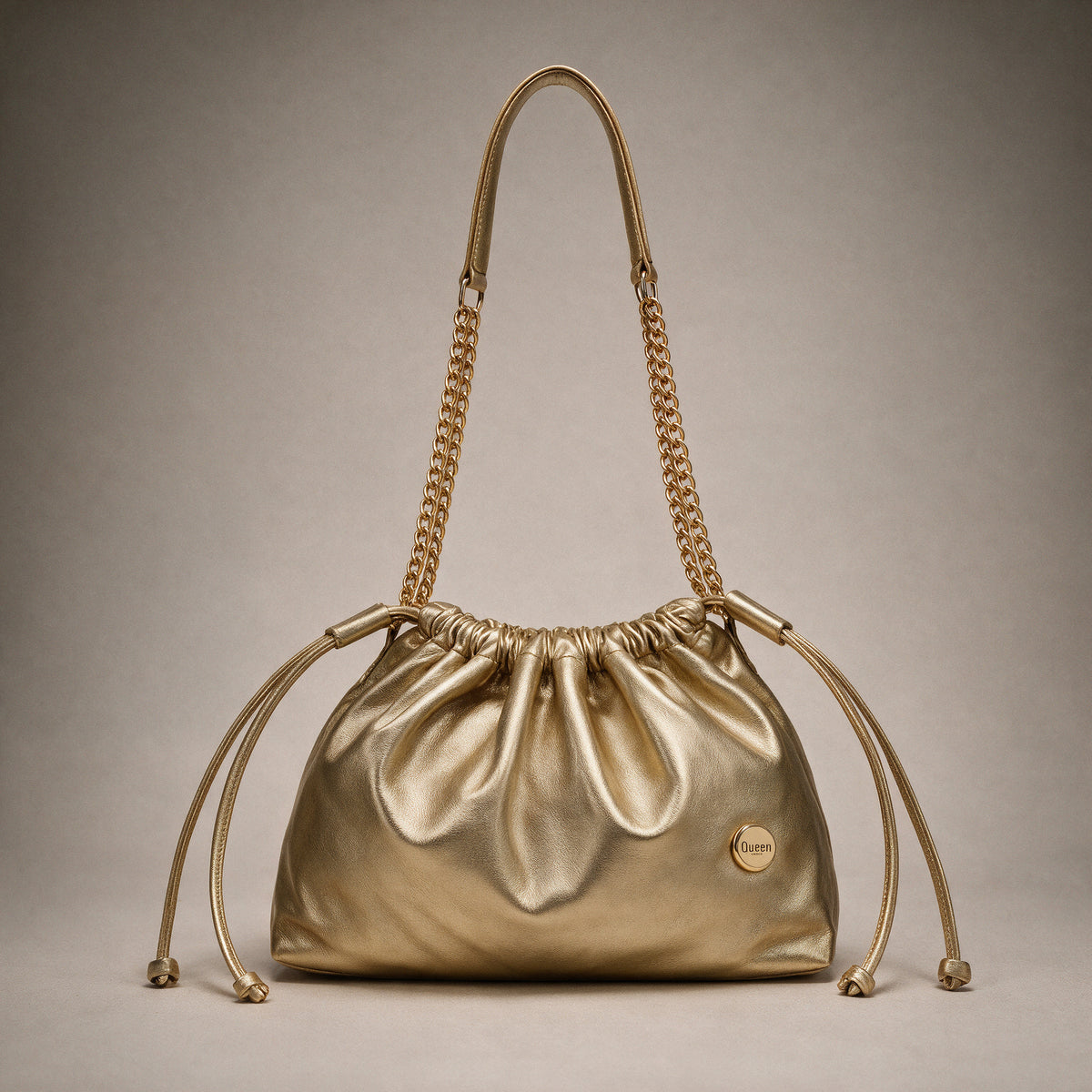 Bolso Mia Oro en cuero dorado metálico visto de frente sobre fondo greige de estudio, con cadena dorada y cordones de cuero