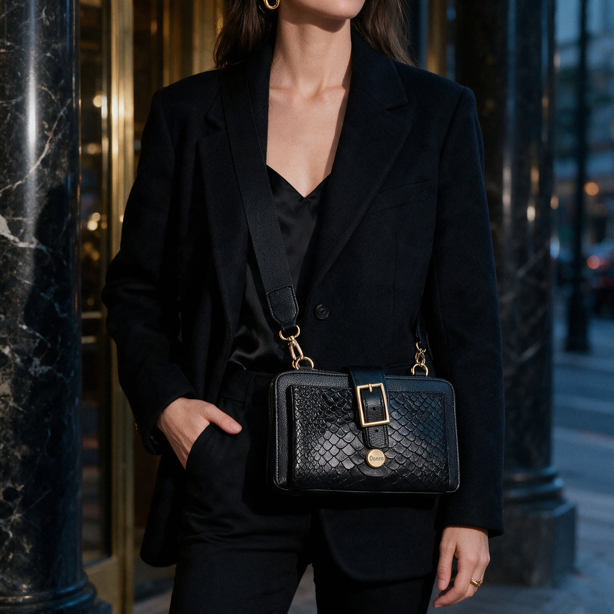 Bolso Charlotte Negro Luxury con hebilla dorada y botón Queen Leather, llevado cruzado por una mujer con blazer negro a la entrada de un edificio elegante
