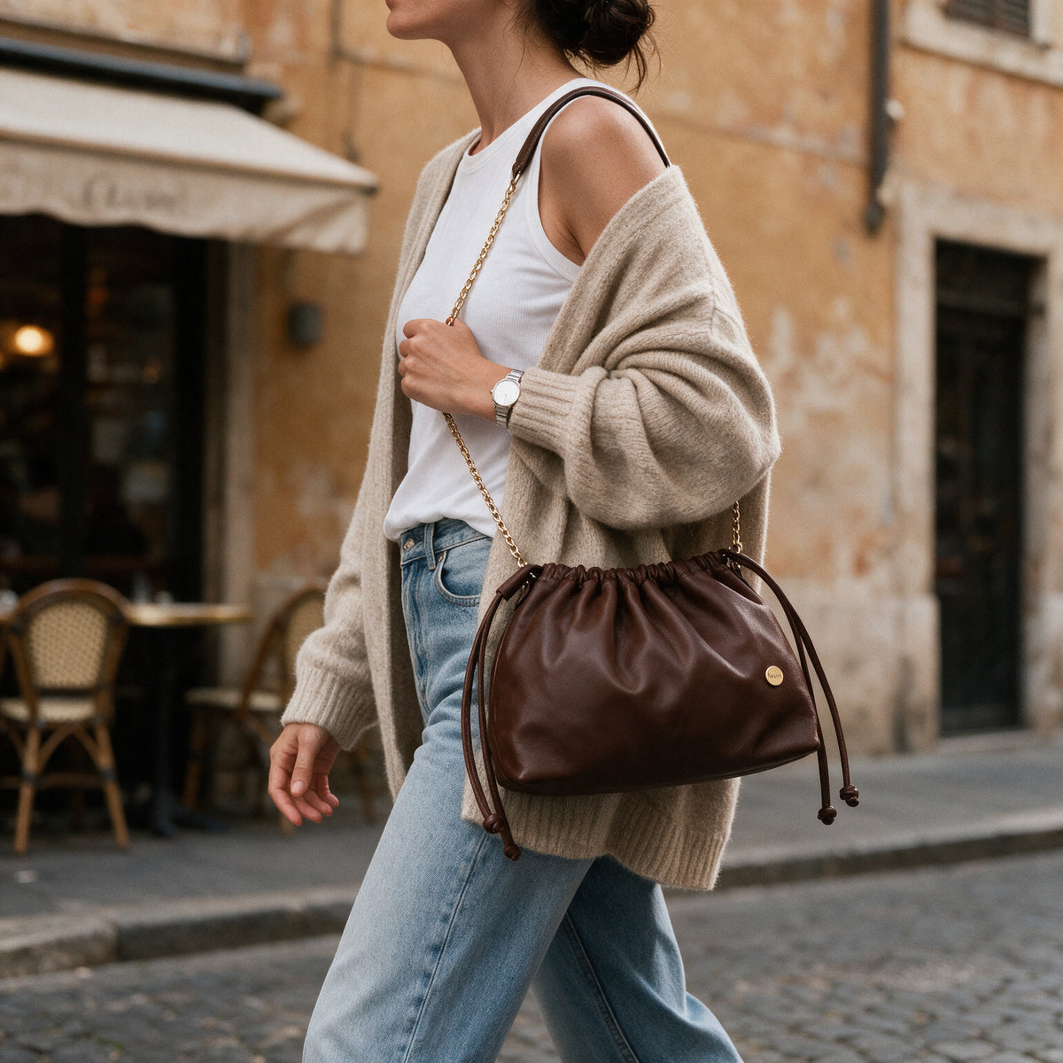 Bolso Mia en cuero chocolate mate con cadena dorada, llevado al hombro por una mujer con jeans, camiseta blanca y cárdigan de lana en una calle empedrada