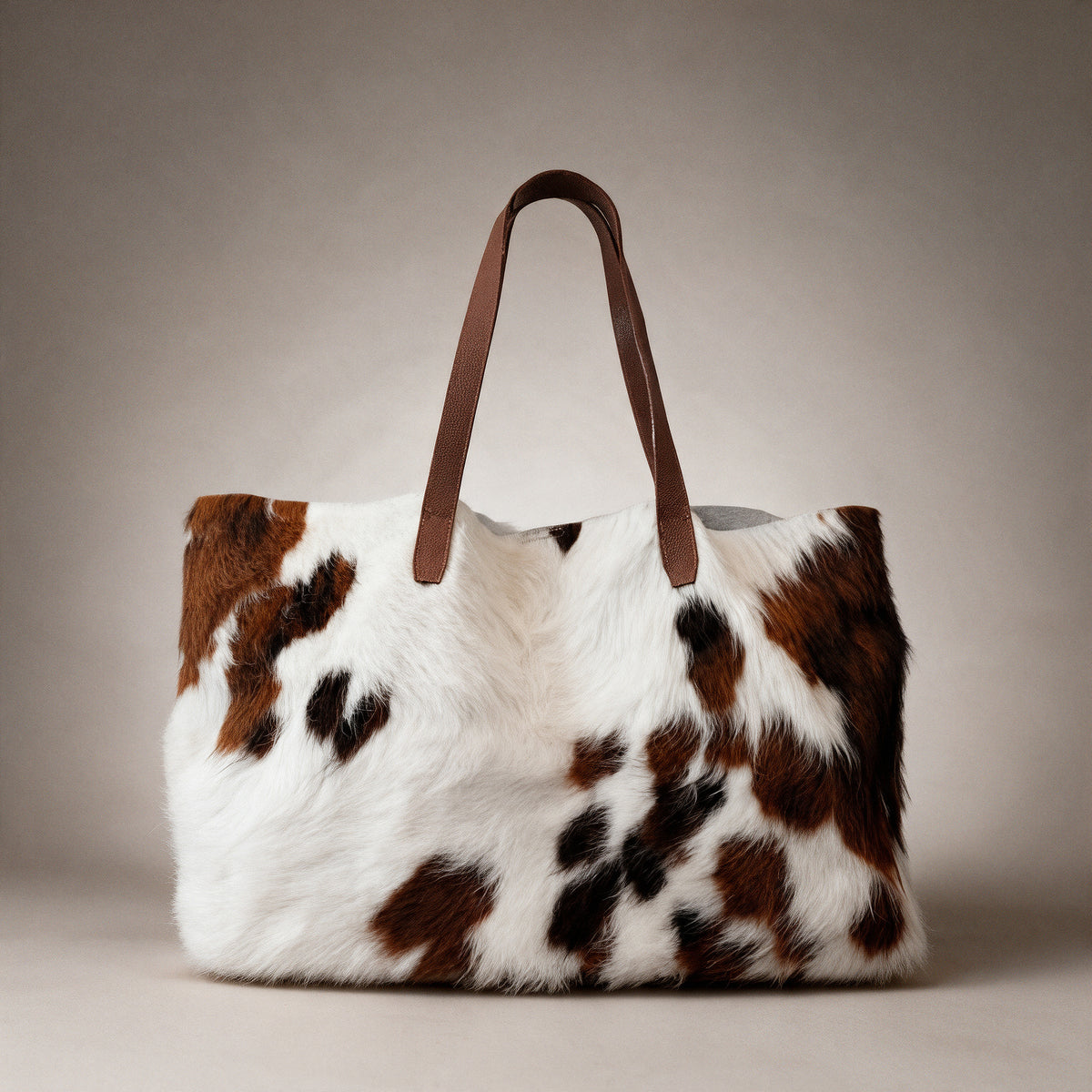 Bolsa tote en pelo natural blanco con manchas marrones vista de frente sobre fondo greige de estudio, con asas de cuero