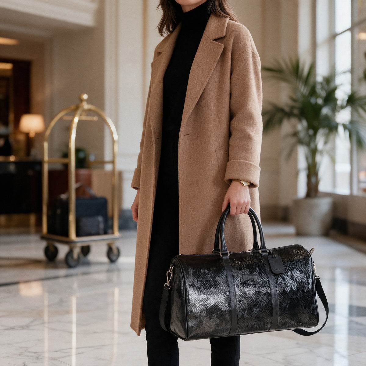 Bolso de viaje Bag Queen Travel Negro en cuero con grabado de serpiente y patrón camuflado, llevado en mano por una mujer con abrigo camel en el lobby de un hotel
